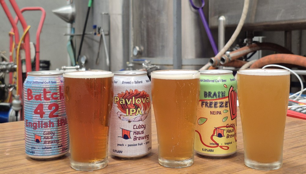 Three IPAs in cans and glasses in front of the Cubby Brew Haus